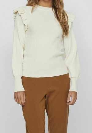 Jersey blanco de manga larga con detalles de volantes en los hombros, combinado con pantalones marrones de talle alto, en una persona de pie contra un fondo liso.