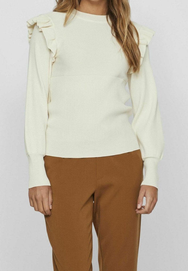 Pull blanc à manches longues avec détails à volants sur les épaules, associé à un pantalon marron taille haute, porté par une personne debout sur un fond uni.