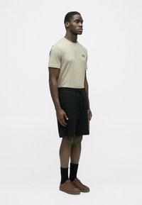 Bézs rövid ujjú póló logóval, fekete rövidnadrággal, amelyen arany oldalnyomat található. A megjelenést barna sportcipő és fekete edzőzokni egészíti ki.