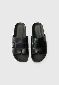 Black slide sandals with a smooth leather finish, featuring cut-out design and a wide strap across the foot. Brand logo embossed on top.