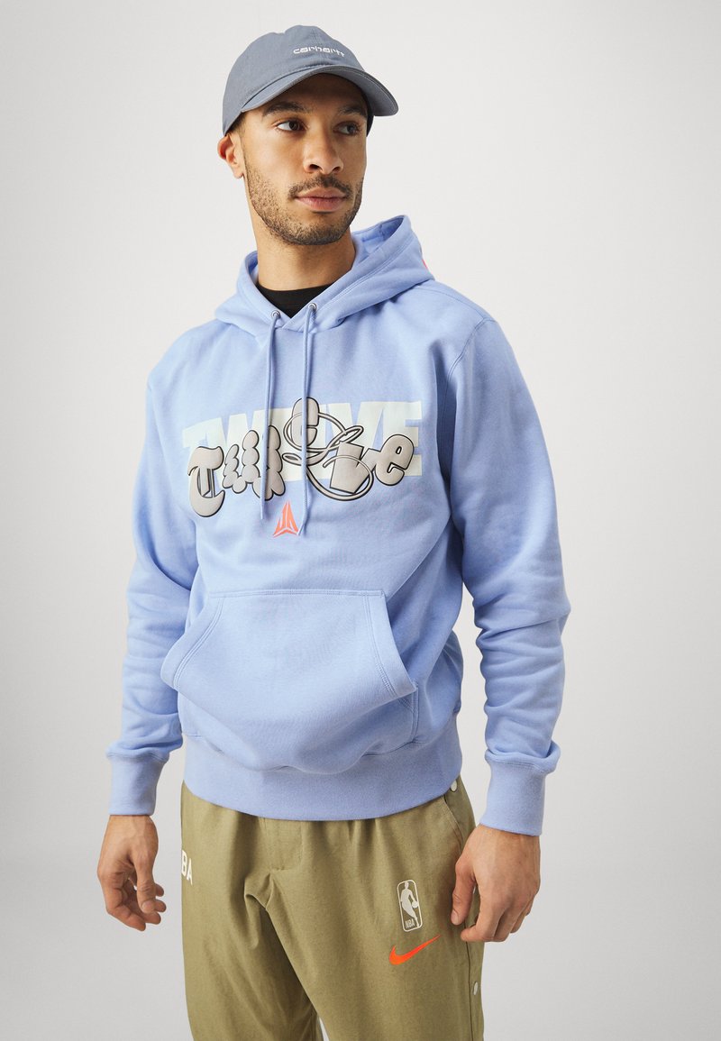 Sudadera azul claro con capucha ajustable y bolsillo frontal. Presenta un destacado diseño gráfico en texto. Combinada con pantalones beige y una gorra gris.