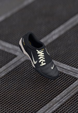 Czarny sneaker Nike z białymi sznurowadłami i logo, unoszący się nad ciemną metalową kratownicą podłogową.