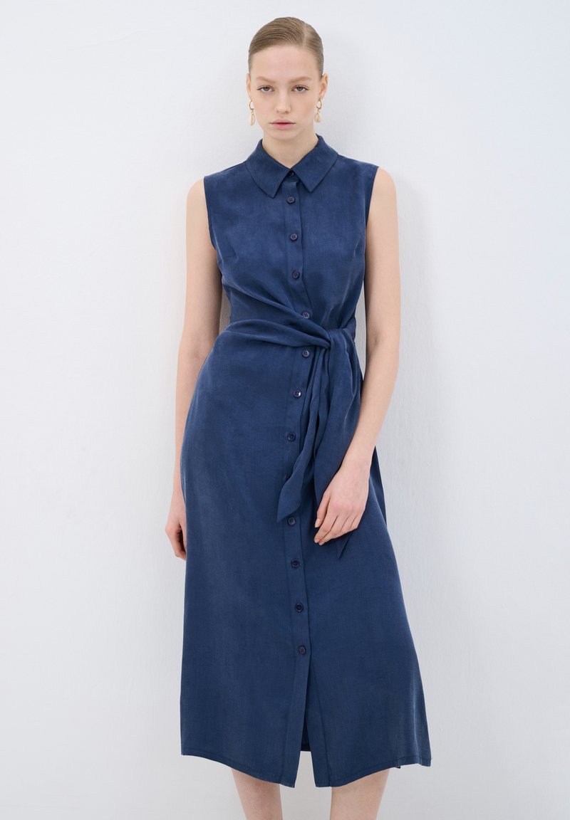 Robe sans manches à boutons en tissu bleu marine avec une taille nouée. Elle présente un col et une longueur droite, offrant une apparence lisse et texturée.