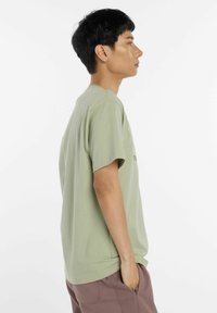 T-shirt en coton vert olive à manches courtes, avec une coupe décontractée et une petite inscription sur le devant. Porté avec un pantalon marron, les mains dans les poches.