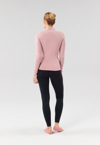 Haut ajusté à manches longues en tissu rose doux, associé à des leggings noirs. Texture lisse sans motifs visibles, design minimaliste.