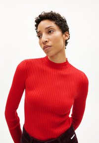 Haut à manches longues rouge côtelé avec un col montant. La texture du tissu est lisse avec des lignes verticales. Porté avec un pantalon de couleur sombre.