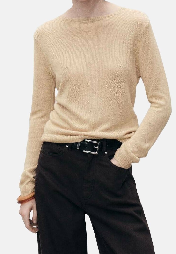 Personne portant un pull beige à manches longues, rentré dans un pantalon noir taille haute avec une ceinture noire et un bracelet marron au poignet gauche.