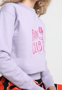 Lila sweatshirt med en grafik i rosa text som säger "FAN CLUB" med en rosillustration, tillverkad av mjukt bomullsmaterial med en avslappnad passform.