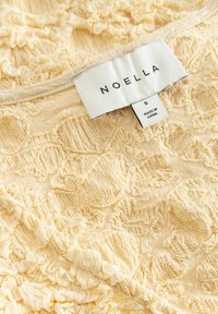 Étiquette de vêtement « NOELLA » taille petite sur tissu jaune pâle texturé avec motif floral en relief et froncé. Étiquette indiquant « Fabriqué en Chine » visible.