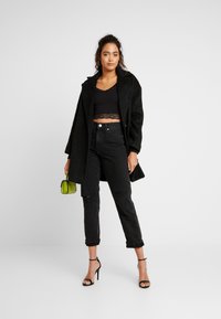 Black wool coat with a structured design, layered over a black lace crop top. Worn with high-waisted black jeans and black heels, holding a green handbag.