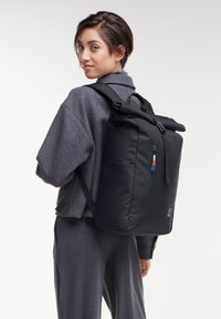 Zaino roll-top nero realizzato in tessuto morbido, dotato di una tasca frontale con zip e tirazip colorati e spallacci regolabili.