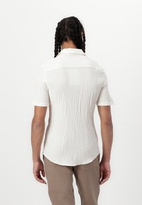 Biała koszula z krótkim rękawem, z fakturowanego materiału, z kołnierzykiem i lekko zaokrąglonym dołem. Gładki materiał z delikatnym marszczeniem na plecach.
