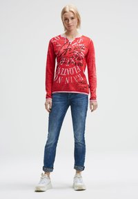 Rotes Langarmhemd mit weißem Kragen, das schwarze Textgrafiken zeigt. Kombiniert mit hochgekrempelten blauen Jeans und weißen Turnschuhen.