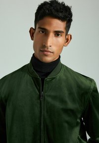 Giacca bomber verde con una texture liscia, colletto a coste e zip frontale; indossata sopra un maglione nero a collo alto.
