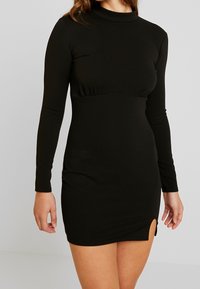 Schwarzes, langärmliges Minikleid aus dehnbarem Stoff. Mit rundem Ausschnitt und seitlichem Schlitz. Glatte Textur mit einer figurbetonten Silhouette.