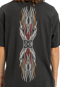 Čierne bavlnené tričko s grafíkami štýlizovaného plameňa v bielej, červenej a oranžovej farbe, umiestnené okolo loga DC na zadnej strane.