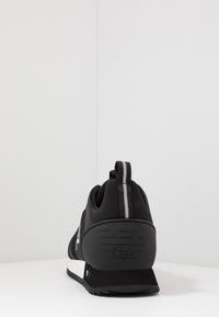 Svart atletisk sko med mesh-övre och gummisula, med en grå randig accent och präglad logotyp på hälen. Bakifrån visad.