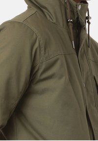 Hombre vestido con una chaqueta verde oliva con un alto cuello, cremallera, botones a presión y cuerdas ajustables en la capucha.