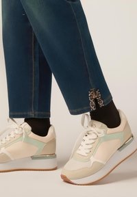 Zapatillas elegantes en blanco y crema con detalles en verde menta, paneles texturizados y una suela blanca elevada. Los jeans presentan detalles metálicos decorativos en el dobladillo.