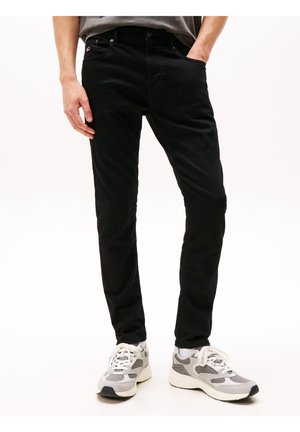 Mand iført slanke sorte jeans og grå og hvide sneakers, stående med den ene hånd i lommen mod en ensfarvet baggrund.