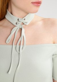 Ljusgrå choker med en slät yta och snörningsdesign som har silveröglor och långa knytningar, bärs med en off-shoulder topp.