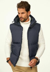 Marineblaue Steppweste mit Reißverschluss vorne, hohem Kragen und Kapuze. Getragen über einem weißen, kabelgestrickten Pullover, mit Seitentaschen und elegantem Design.