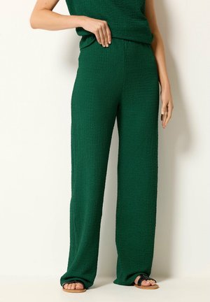 Femme portant un pantalon large texturé vert foncé, associé à un haut sans manches assorti et des sandales plates noires.