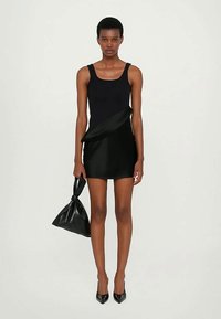 Vestido mini preto com uma silhueta ajustada, apresentando uma textura suave e um detalhe drapeado. Modelo segura uma bolsa preta e usa sapatos de salto agudo.