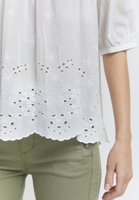 Blouse en coton blanc avec des broderies et des découpes le long de l'ourlet, avec une bordure en forme de vague. Associée à un pantalon ajusté vert olive.