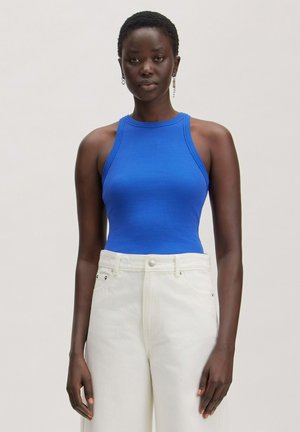 Vrouw met kort haar, die een mouwloos blauw topje en een witte broek met hoge taille draagt, staat tegen een effen lichte achtergrond.