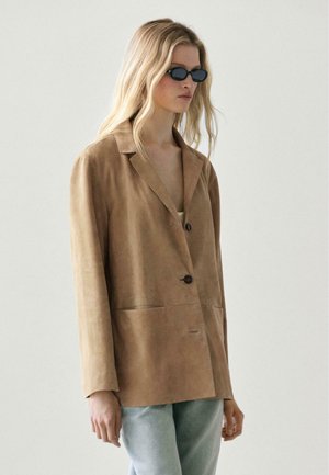 Donna bionda che indossa occhiali da sole ovali neri, blazer oversize in suede color cuoio allacciato con un bottone e jeans azzurri, in piedi su uno sfondo semplice.