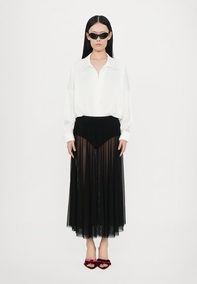 White blouse with a collar, paired with a sheer black skirt featuring pleats and a solid waist. Black sunglasses and red velvet sandals complete the look.