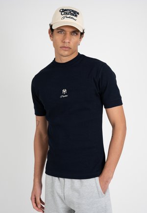 Joven con gorra beige con la inscripción "Champagne Courtside Prohibited" y camiseta negra de manga corta con un emblema blanco, de pie frente a un fondo blanco.