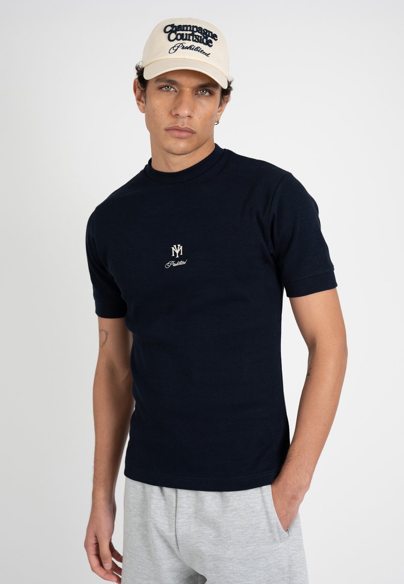 Jeune homme portant une casquette beige avec l'inscription « Champagne Courtside Prohibited » et un t-shirt noir à manches courtes avec un emblème blanc, debout devant un fond blanc.