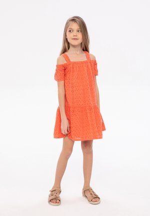 Robe orange sans manches avec des manches bouffantes dénudées, présentant des motifs œillets et un bas évasé, associée à des sandales marron.