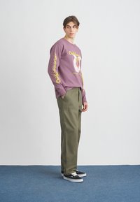 Μακρυμάνικη λεβαντί μπλούζα με κίτρινο γραφικό και κείμενο, συνδυασμένη με παντελόνι ελιάς και μαύρα αθλητικά παπούτσια με λευκές σόλες.