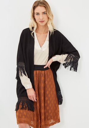 Mujer con cabello rubio que lleva un chal negro con flecos, blusa crema y falda midi estampada en tonos naranjas sobre un fondo claro.