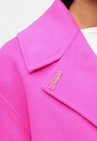 Skaisti rozā mēteli ar zelta kursīvu "N nākotne" piespraudi, kas piesprausta tuvu apkaklei, valkā pār baltu blūzi.