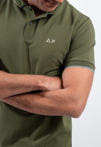 Polo verde oliva con colletto, caratterizzato da un piccolo emblema e dettagli blu sulle maniche. Tessuto in cotone morbido e testurizzato.