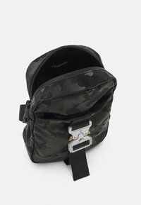 Zaino camouflage nero con superficie testurizzata, dotato di chiusura a fibbia argentea e tracolla regolabile. Scomparto principale con zip.