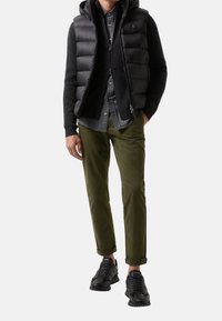 Schwarze Steppweste über einem dunklen Pullover, kariertem Hemd, olivgrünen Hosen und schwarzen Sportschuhen. Merkmale sind Reißverschlüsse und umgeschlagene Ärmel.