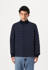 Manteau rembourré marine avec col montant, design matelassé et poignets côtelés. Dispose d'une fermeture éclair à l'avant et de poches latérales.