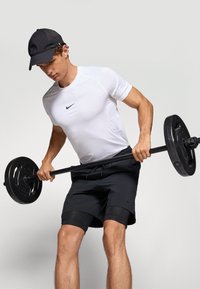Weißes, feuchtigkeitsableitendes Sportshirt mit schwarzem Logo, kombiniert mit schwarzen Shorts, während er eine schwarze Langhantel mit Gewichtsscheiben hält.