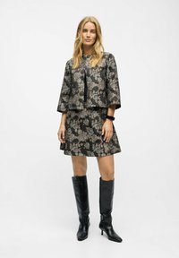 Kvinde iført en sort blomstret jakke og nederdel med sorte knæhøje hæle mod en plain hvid baggrund.