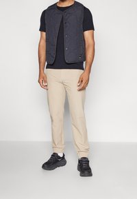 Černá prošívaná vesta přes černé tričko, doplněná béžovými tapered kalhotami a černými teniskami s bílými akcenty. Hladké textury látek.