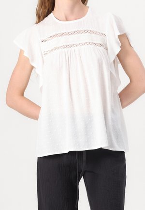 Femme portant un chemisier blanc texturé avec des inserts en dentelle et des manches volantées, associé à un pantalon noir ample avec une taille à cordon.
