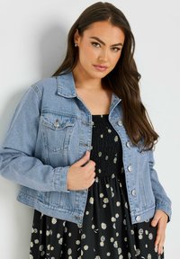 Giacca di jeans azzurro chiaro con colletto, tasche frontali e bottoni argentati, indossata sopra un vestito nero con fantasie floreali bianche.