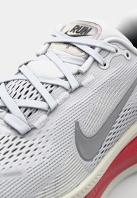Baskets de sport gris clair avec tige en maillage, accents réfléchissants et lacets visibles. Présente un swoosh Nike gris et une semelle extérieure rouge contrastante.
