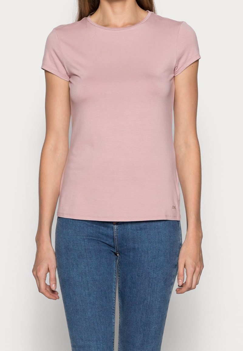 Femme portant un t-shirt ajusté rose clair à manches courtes et un jean bleu, debout devant un fond clair uni.