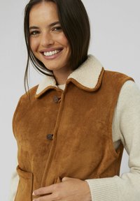 Gilet en daim marron avec un col crème doux, doté de deux poches avant et de boutons marron, porté sur un pull léger.